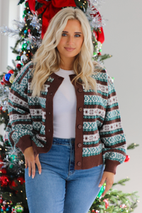Close up of a woman model wearing a brown, green, and white Christmas themed sweater with multiple button closures in the front, slightly puffed sleeves, and a relaxed fit. The cardigan is paired with a white top underneath and medium wash denim jeans. The photo is taken inside a modern home with a decorated Christmas tree in the back. 