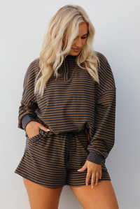 Close up of a woman model wearing a brown and multi-colored striped pattern throughout. The set consists of an oversized top with quarter-button details and shorts. The photo is taken outdoors against a white wall. 