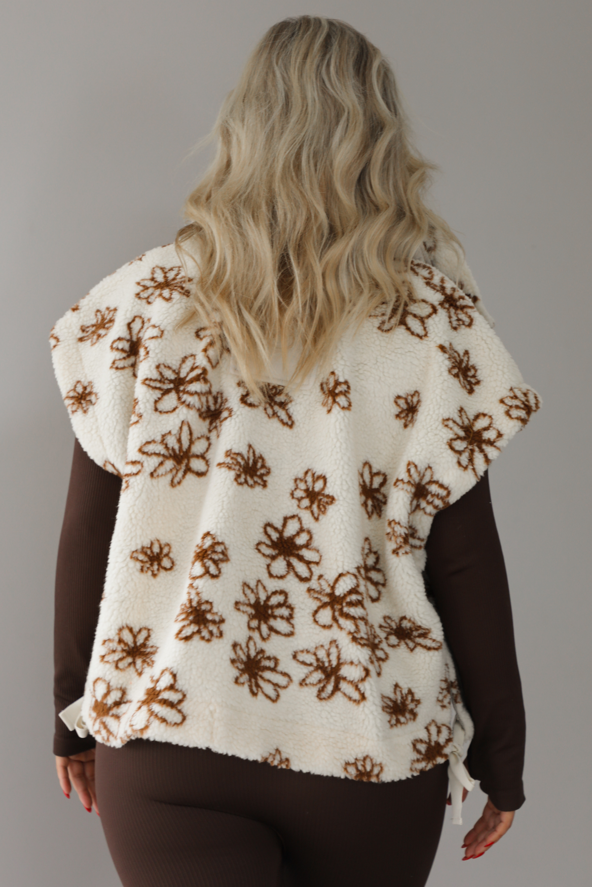 The photo features a model wearing a beige sherpa vest with a brown floral print pattern throughout, functional button closures in the front, and that is oversized with a brown, fitted jumpsuit. The photo is taken indoors against a white wall with white marbled flooring. The photo shows the back of the vest.