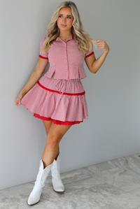 Woman wearing a red and white gingham patterned set that has a short sleeved button-up top that has pleated details at the hem of the top and a high waisted skirt with red lace detailing at the hem. The set is paired with white cowboy boots. The photo is taken indoors against a white wall with white marble flooring. 