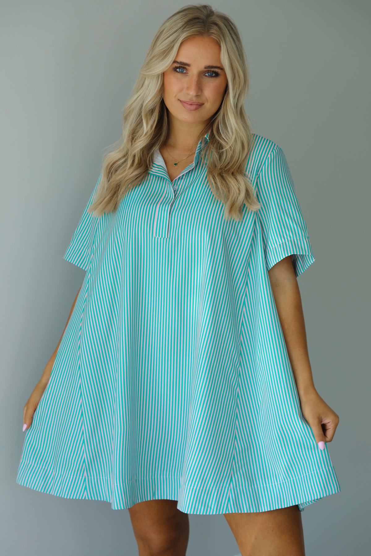 Woman wearing a green and pink striped dress that has a collared neckline, functional quarter button closures in the front, and a flowy skirt. The photo is taken indoors against a white wall.