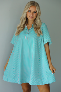 Woman wearing a green and pink striped dress that has a collared neckline, functional quarter button closures in the front, and a flowy skirt. The photo is taken indoors against a white wall.