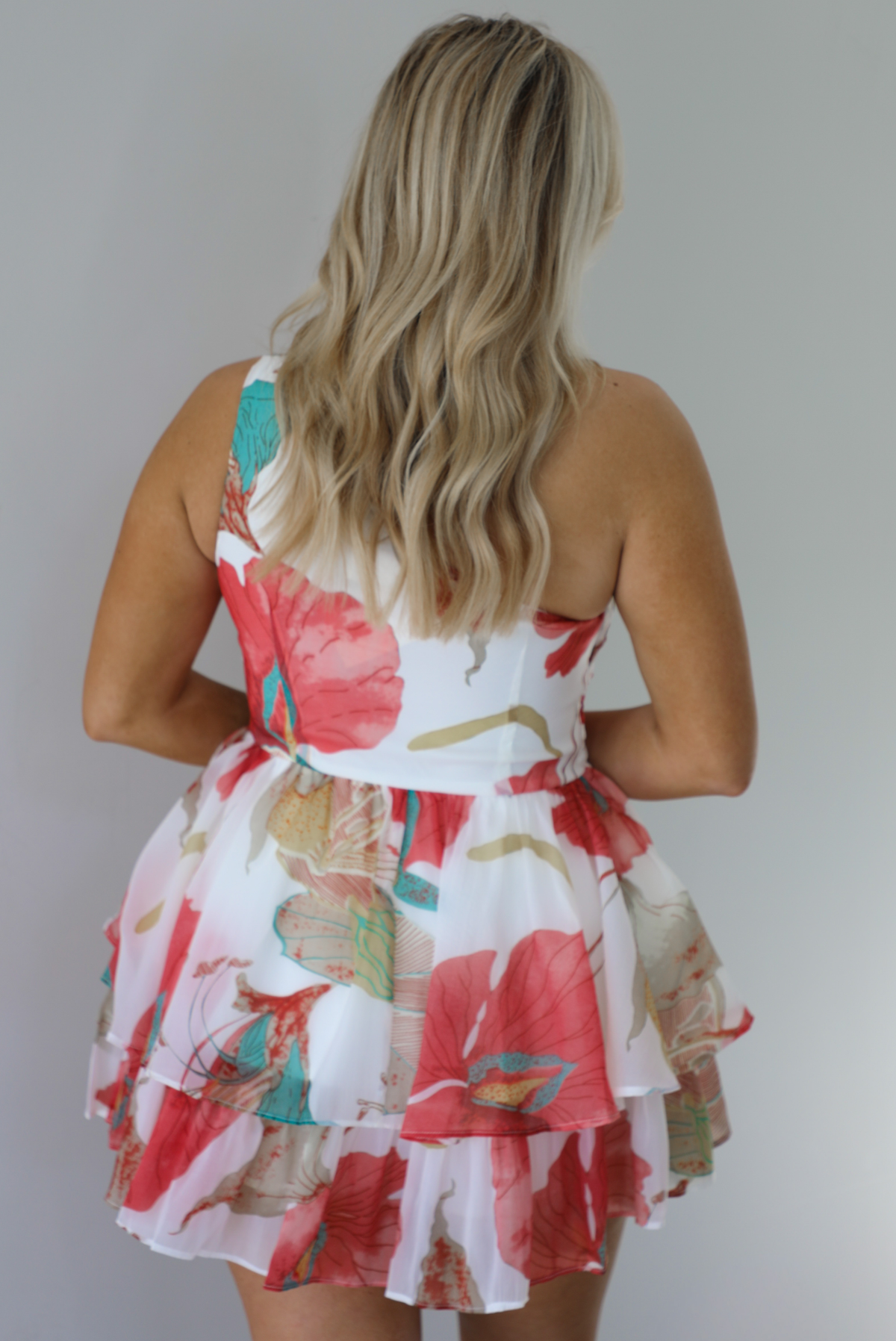Woman wearing a one strap white and multi-colored floral dress with a ruffled skirt. The photo is taken indoors against a white wall and shows the back of the romper. 