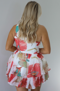 Woman wearing a one strap white and multi-colored floral dress with a ruffled skirt. The photo is taken indoors against a white wall and shows the back of the romper. 