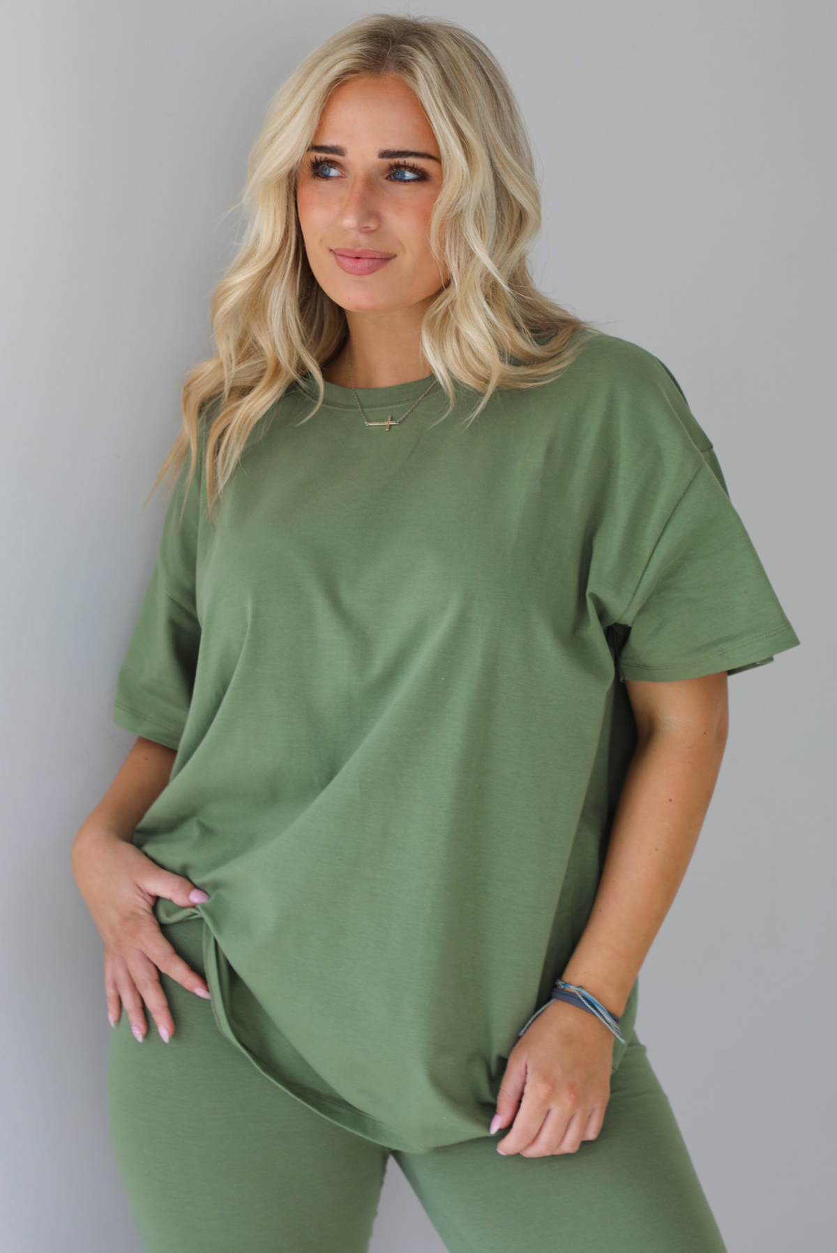 Woman wearing a green two piece set featuring an oversized t-shirt and fitted leggings. The photo shows a closeup of the oversized top. The photo is taken indoors against a white wall.