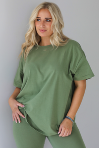Woman wearing a green two piece set featuring an oversized t-shirt and fitted leggings. The photo shows a closeup of the oversized top. The photo is taken indoors against a white wall.