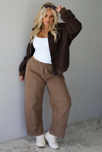 A woman wearing a dark brown suede jacket with a white tank and mocha colored mid-to-low rise pants, standing indoors against a white wall with white marble flooring.
