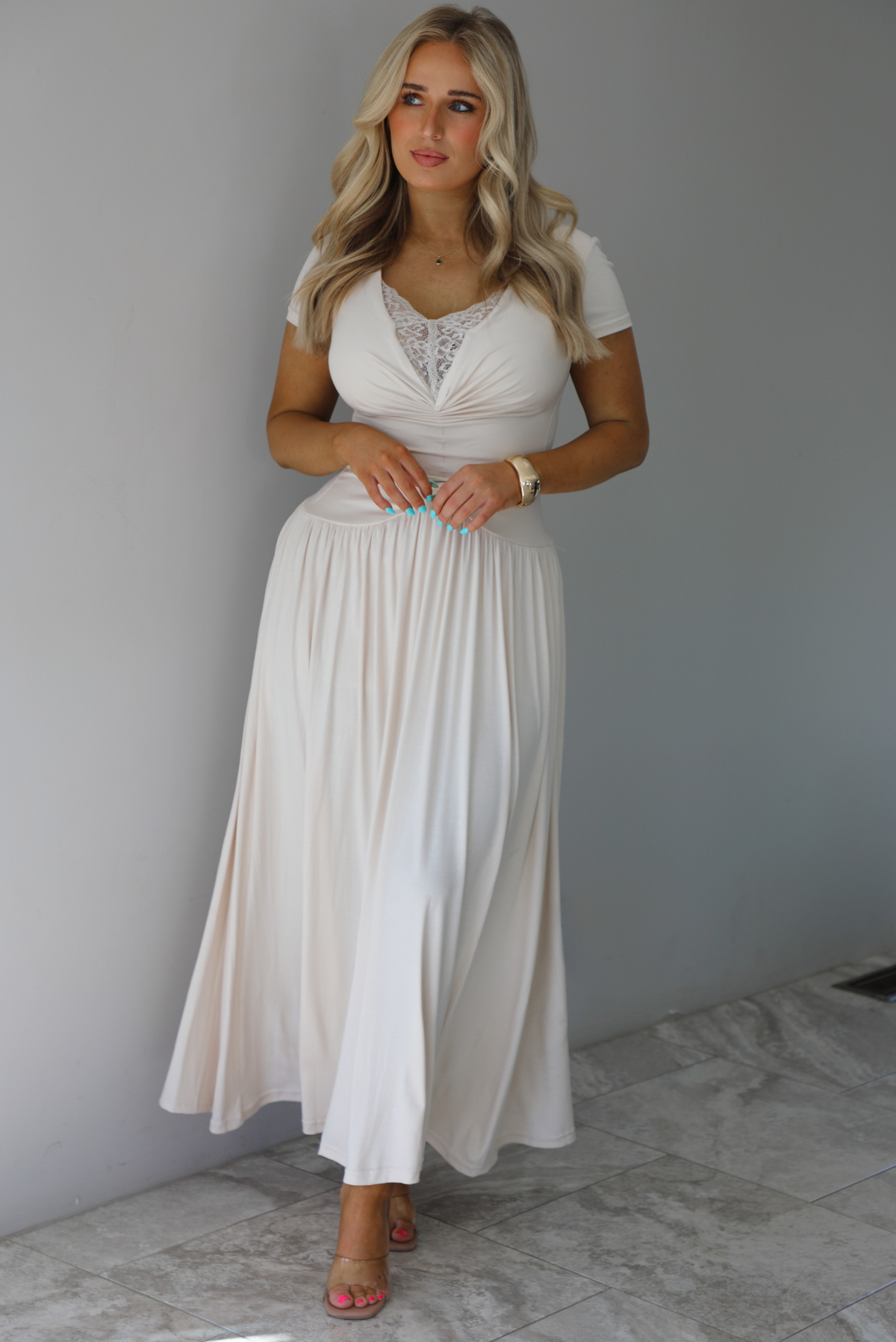 Woman wearing a light sand colored maxi length dress that has a fitted bodice with lace detailing in the center of the bust, a drop waist design, short sleeves, and a flowy skirt. The photo is taken indoors against a white wall with white marble flooring. 