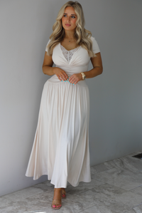 Woman wearing a light sand colored maxi length dress that has a fitted bodice with lace detailing in the center of the bust, a drop waist design, short sleeves, and a flowy skirt. The photo is taken indoors against a white wall with white marble flooring. 