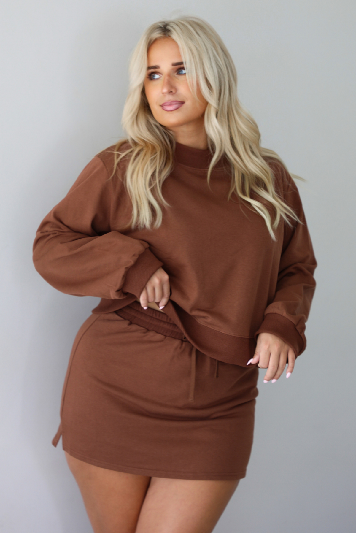 A woman wearing a brown matching two piece set featuring a cropped sweatshirt and mini skirt of the same material, standing indoors against a white wall. The model tucked the top to show the full length. 