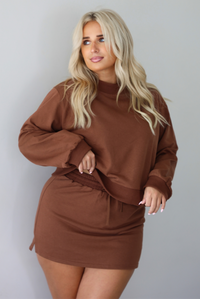 A woman wearing a brown matching two piece set featuring a cropped sweatshirt and mini skirt of the same material, standing indoors against a white wall. The model tucked the top to show the full length. 