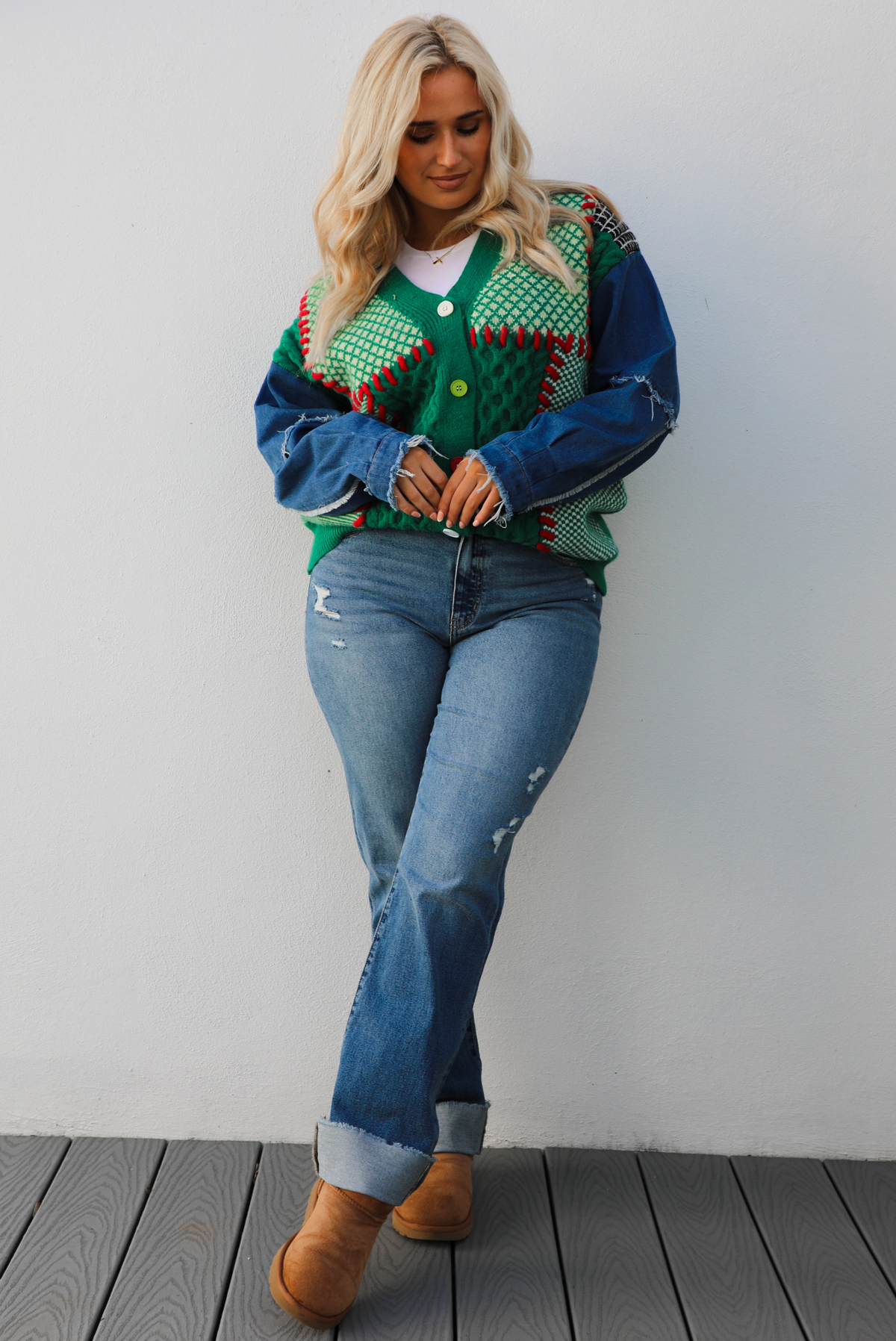 Woman model wearing a green and multi-colored cardigan with denim sleeves, medium wash denim jeans, and tan uggs. The photo is taken against a white wall outdoors.