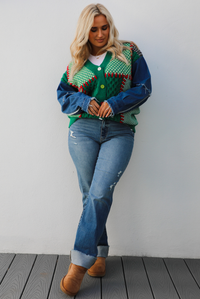 Woman model wearing a green and multi-colored cardigan with denim sleeves, medium wash denim jeans, and tan uggs. The photo is taken against a white wall outdoors.