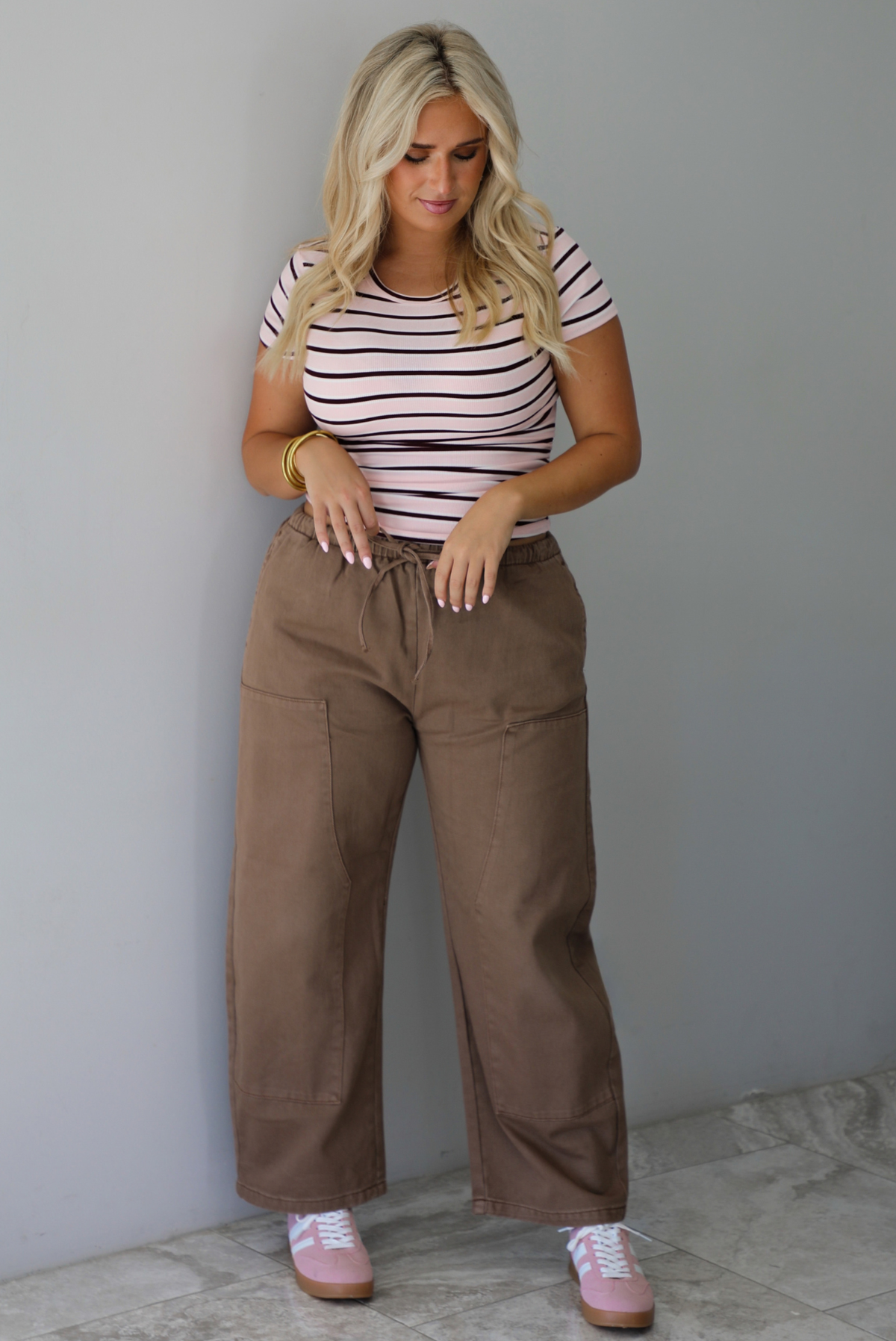A woman wearing a pink baby tee that has a burgundy and white striped design throughout, paired with mocha barrel denim, standing indoors against a white wall with white marble flooring. 