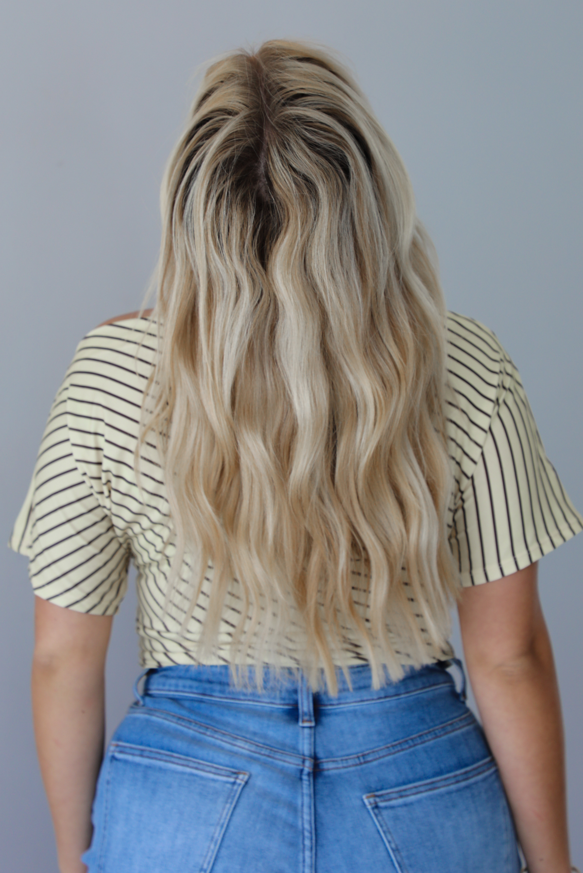 Woman wearing a pale yellow top with a taupe striped pattern with medium wash denim shorts indoors against a white wall. The photo shows the back of the top. 