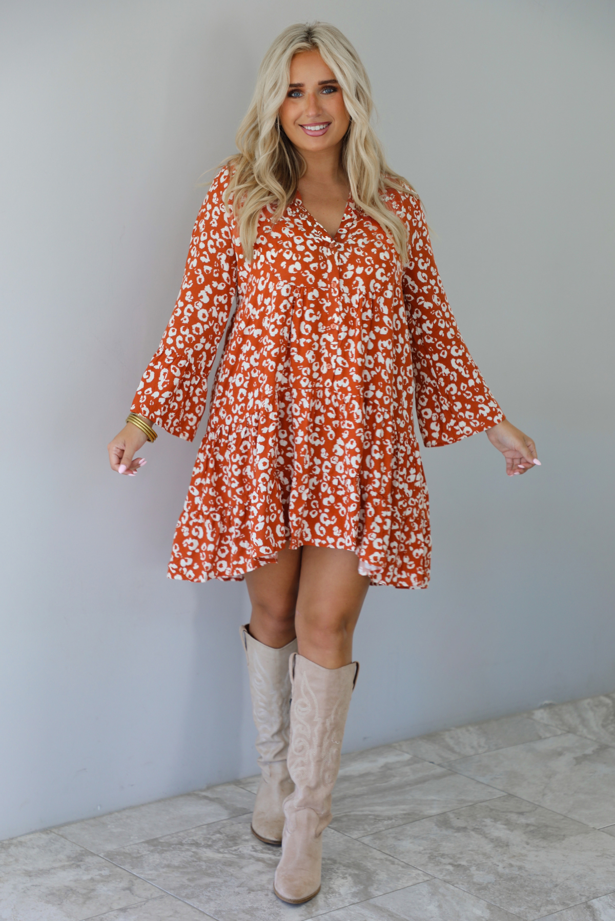 A woman wearing an orange and white animal print long sleeved, flowy mini dress, standing indoors against a white wall with white marble flooring. 