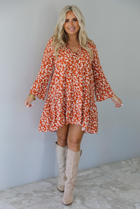 A woman wearing an orange and white animal print long sleeved, flowy mini dress, standing indoors against a white wall with white marble flooring. 
