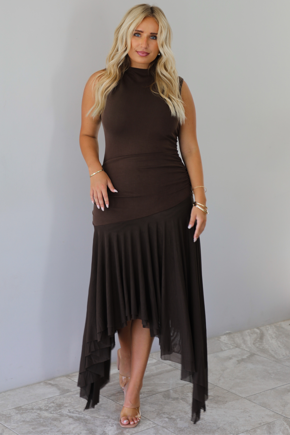 A woman wearing a brown midi dress that has a high neckline, is fitted until the upper thigh, then leads to sheer mesh that's flowy ands an asymmetrical hem, standing indoors against a white wall with white marble flooring. 