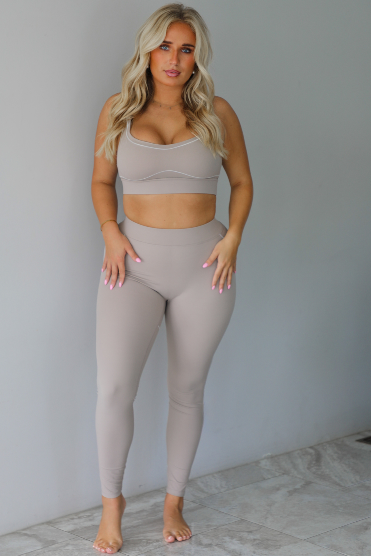 A woman modeling a matching tan activewear set consisting of a sports bra and high-waisted leggings with white stitching details, standing indoors against a white wall with white marble flooring. 