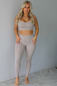 A woman modeling a matching tan activewear set consisting of a sports bra and high-waisted leggings with white stitching details, standing indoors against a white wall with white marble flooring. 