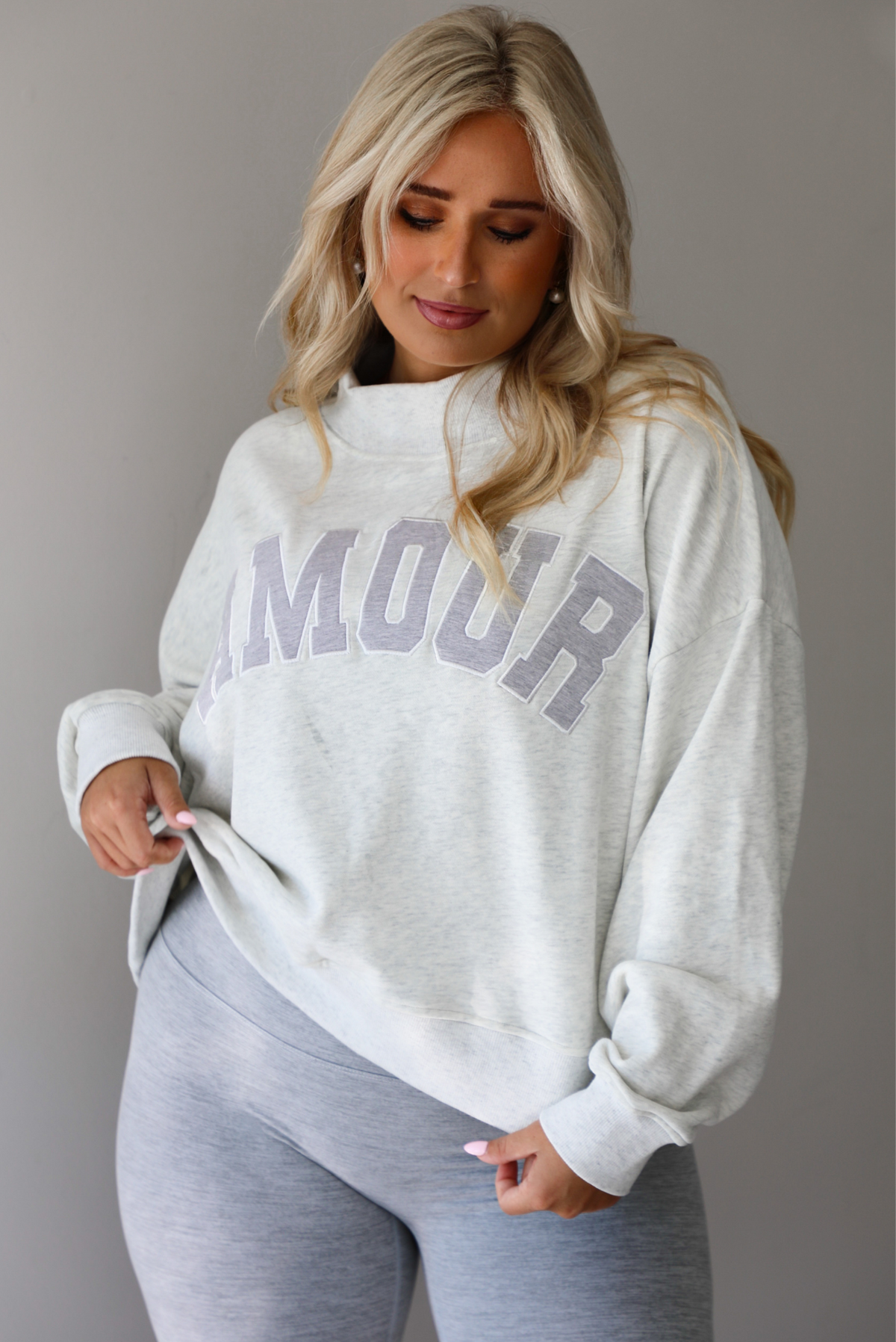 A woman posing in a light gray sweatshirt with 'AMOUR' printed in gray letters and light heather gray leggings, standing indoors against a white wall. The photo shows a closeup of the sweatshirt, showing how it has a high mock neck and is oversized. 