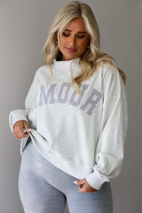 A woman posing in a light gray sweatshirt with 'AMOUR' printed in gray letters and light heather gray leggings, standing indoors against a white wall. The photo shows a closeup of the sweatshirt, showing how it has a high mock neck and is oversized. 