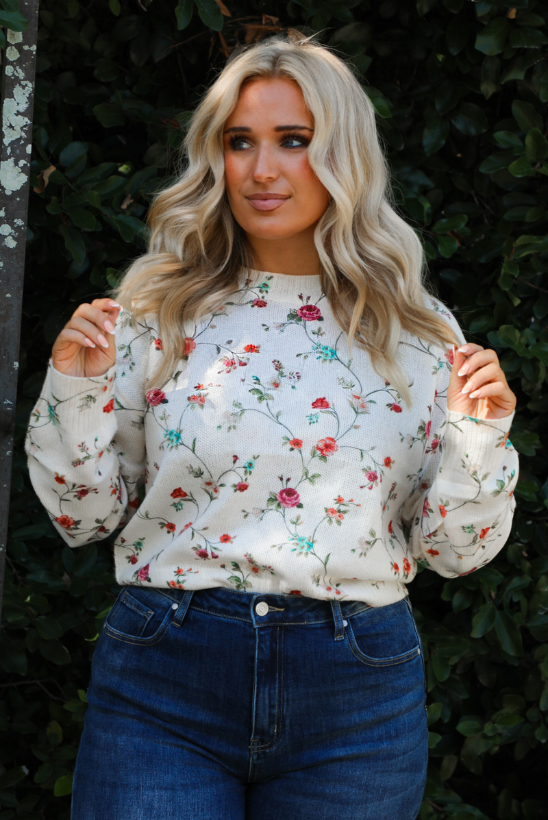 Woman model wearing an off-white sweater that has a multi-colored floral print pattern throughout with dark denim jeans. The photo showcases the length of the sweater and is close up with a slight tuck in the front of the jeans. The photo is taken next to an iron gate outdoors with greenery in the back. 
