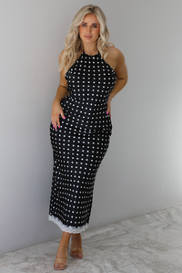 Woman wearing a black wearing a white polka dot printed midi dress that has a halter tie neckline, is form fitting, with white lace trim at the bottom. The photo is taken indoors against a white wall with white marble flooring. 