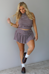 A woman wearing a burgundy and grey striped two piece set featuring a high-neck cropped top and mini skirt, high scrunched white socks, and brown leather loafers, standing indoors against a white wall with white marble flooring. 