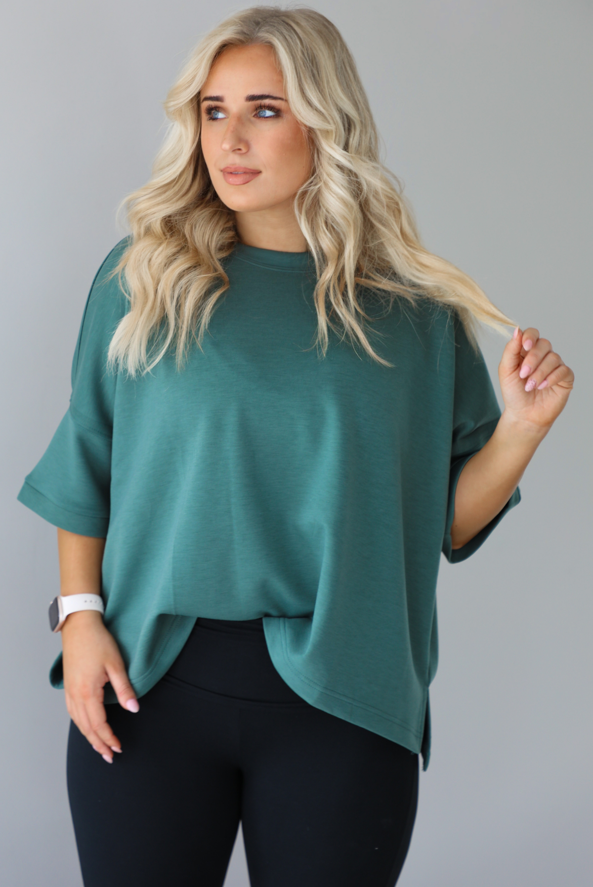 Woman in a teal oversized t-shirt paired with black leggings. The photo shows a closeup of the top which is tucked into the leggings in the front.The photo is taken indoors against a white wall.