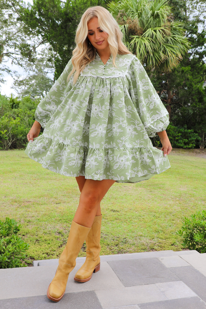 Woman model wearing a green dress that has a checkered and white embroidered floral print pattern throughout. The dress has a babydoll cut style with long sleeves and a flowy fit. It's paired with yellow tall boots. The photo is taken outdoors with grass and trees in the back.