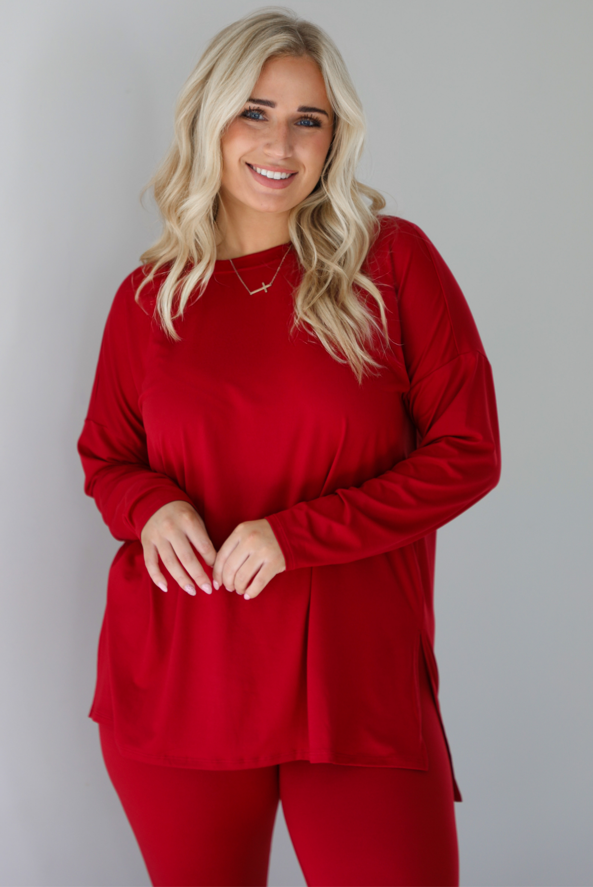 Woman wearing a red two piece set that features an oversized long sleeved top and fitted leggings. The photo shows a closeup of the top and shows how it has split hem details on either side. The photo is taken indoors against a white wall.