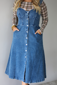 A woman wearing a medium wash denim midi dress with button closures along the  front, with pockets on either side, paired with a sheer, multi-colored plaid long sleeved top underneath, standing indoors against a white wall. The photo shows a closeup of the dress without the models face, showing the models hands in pockets on either side. 