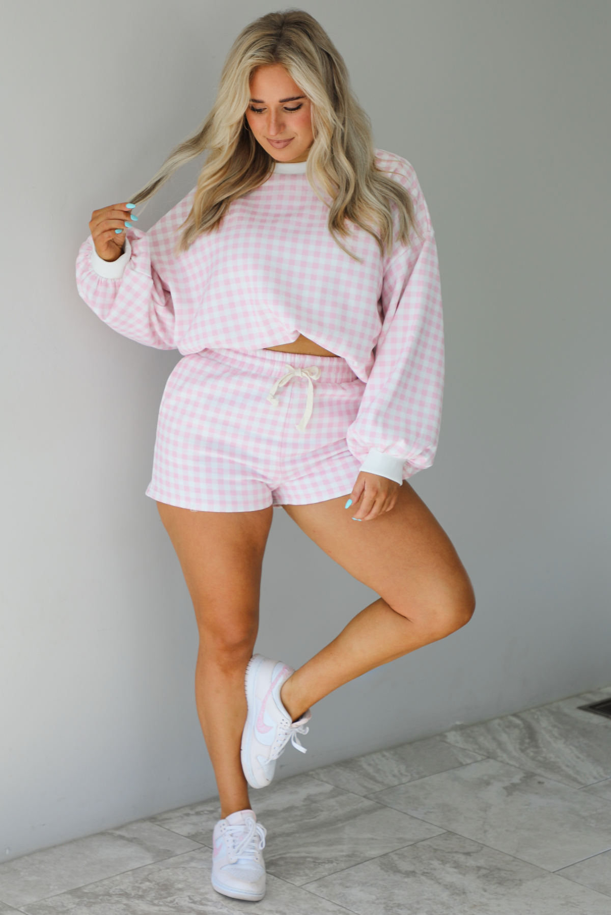 Woman wearing a pink and white checkered print two piece set featuring a sweatshirt and high waisted shorts. The photo is taken indoors against a white wall with white marble flooring. 