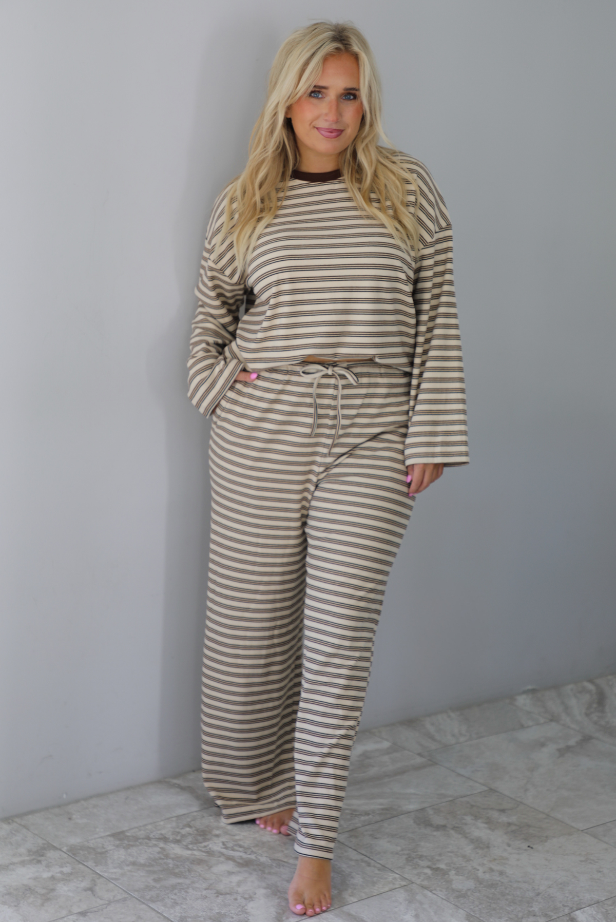 A woman wearing a sand colored matching two piece set with a brown striped pattern throughout featuring an oversized long sleeved top and oversized slouchy wide leg pants, standing indoors against a white wall with white marble flooring. 