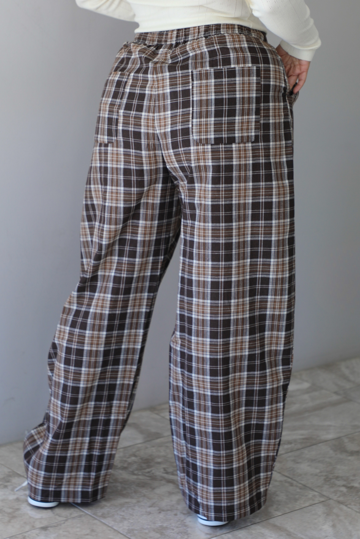 A woman shown from the waist down and from behind wearing a cream colored long sleeved top paired with brown plaid pants, standing indoors against a white wall with white marble flooring. 