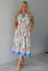 A woman wearing a short sleeved midi dress with a colorful tropical leaf print and light blue trim indoors against a white wall with white marble flooring. 