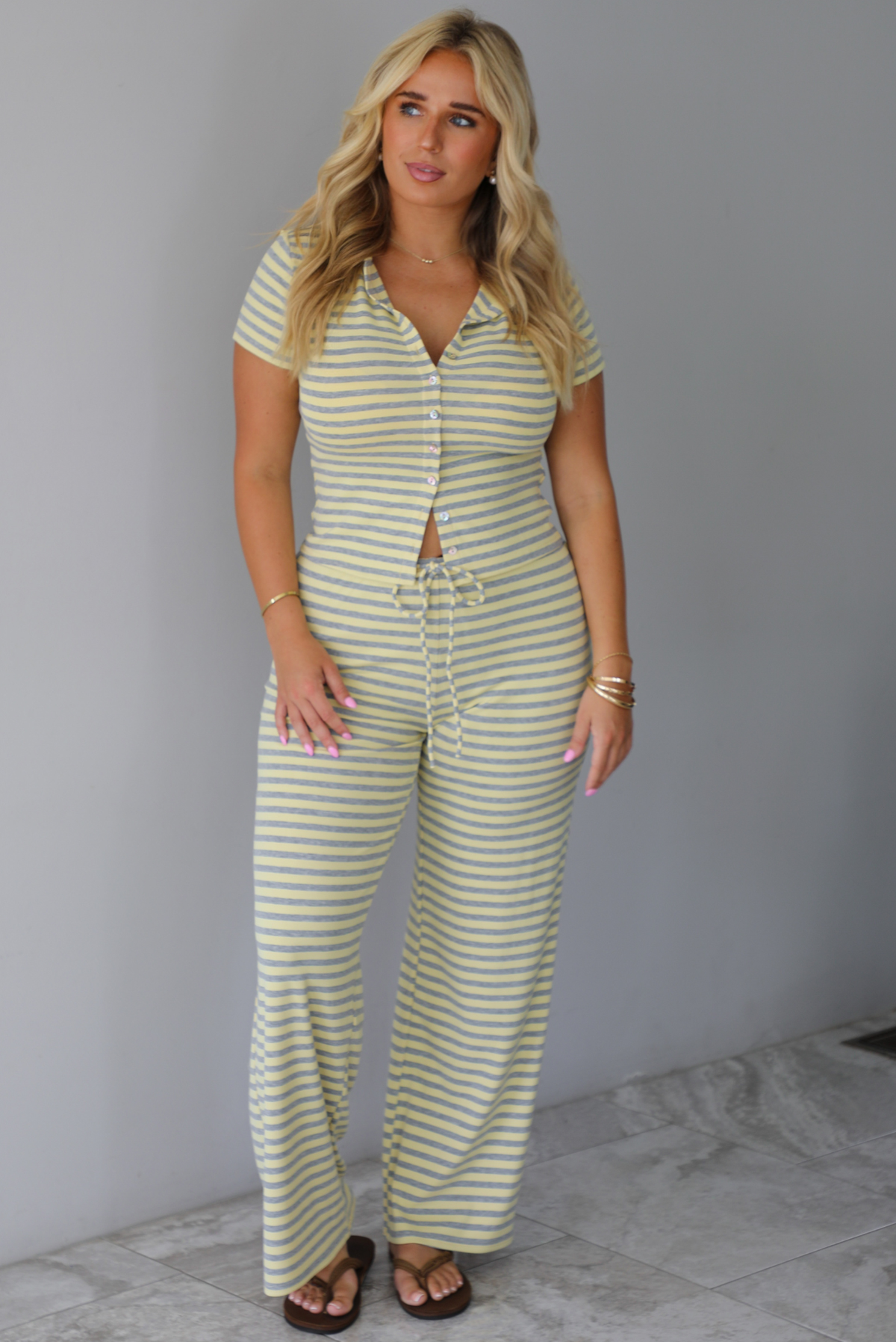 A woman wearing a two piece set consisting of a collared, button-down, short sleeve top and high waisted, drawstring, wide leg pants, standing indoors against a white wall with white marble flooring. 