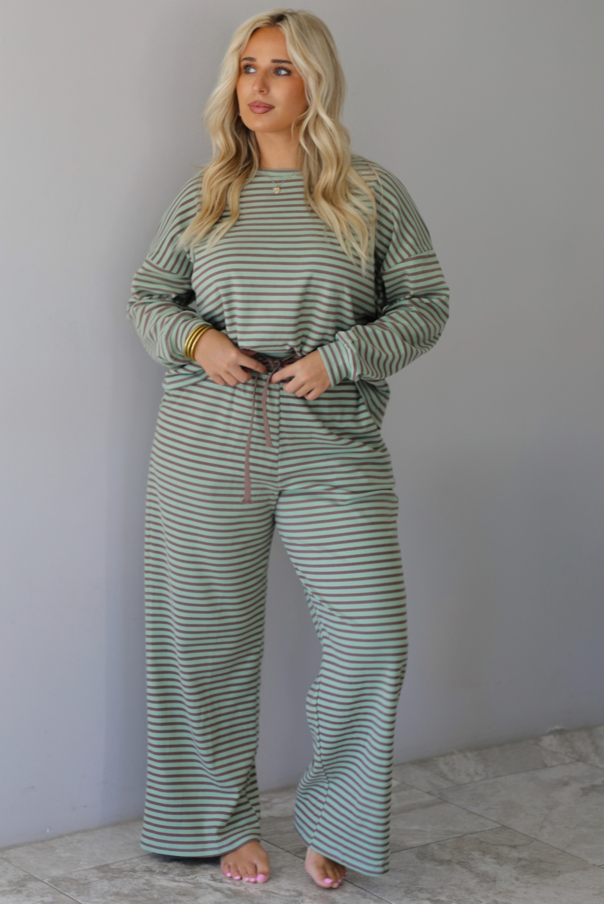 A woman wearing a pale green and brown striped two piece set featuring an oversized long sleeved top and high waisted, wide leg pants, standing indoors against a white wall with white marble flooring. 