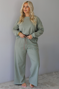 A woman wearing a pale green and brown striped two piece set featuring an oversized long sleeved top and high waisted, wide leg pants, standing indoors against a white wall with white marble flooring. 