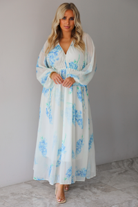 A woman modeling a white maxi dress with a blue floral print and a sheer, flowing overlay, standing indoors against a white wall with white marble flooring. 