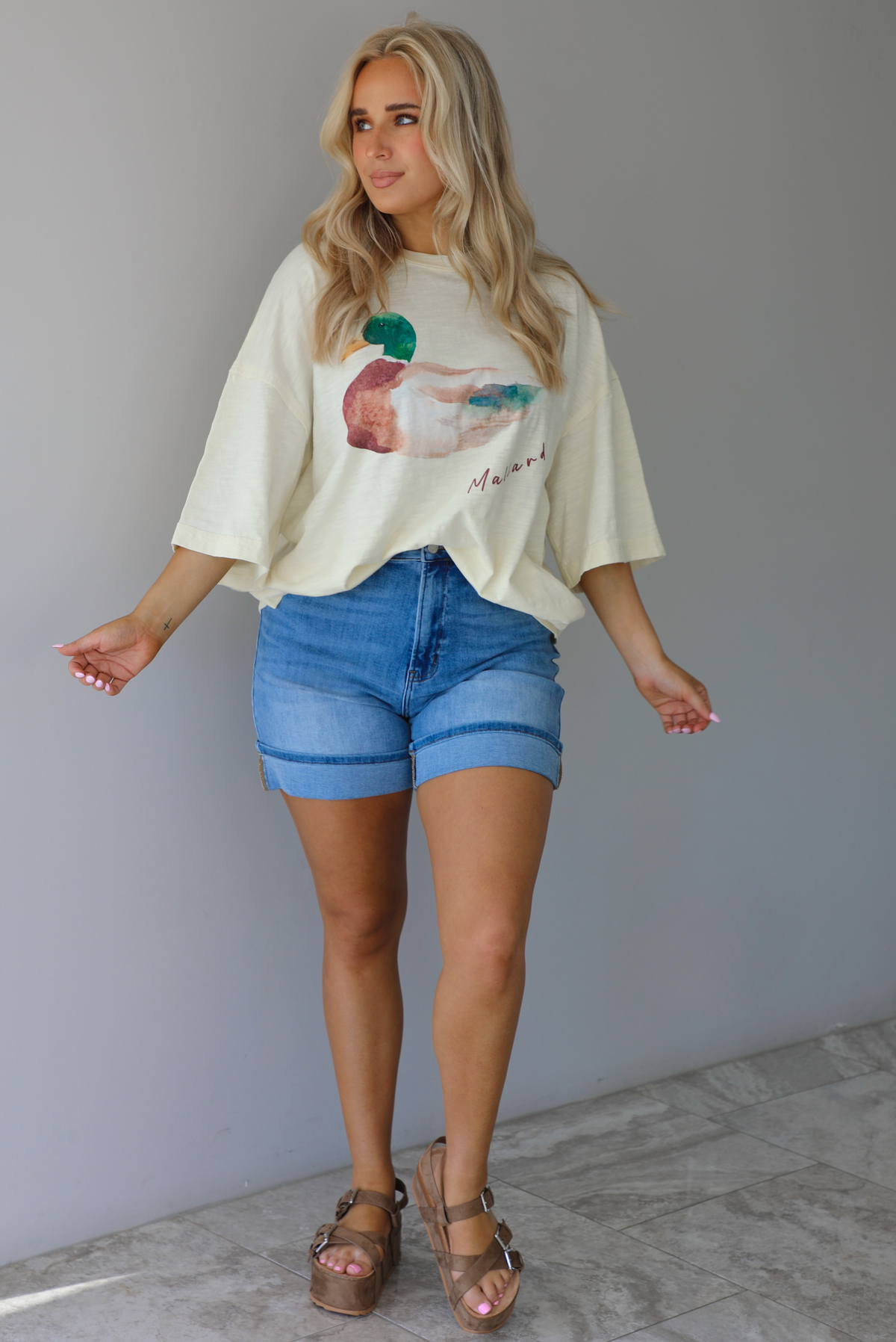 Woman wearing an off-white t-shirt with a graphic design of a mallard duck on the front cuffed into blue denim shorts. The photo is taken indoors against a white wall with white marble flooring. 