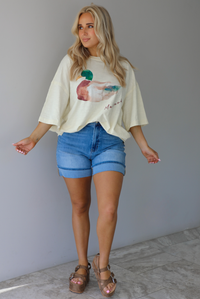 Woman wearing an off-white t-shirt with a graphic design of a mallard duck on the front cuffed into blue denim shorts. The photo is taken indoors against a white wall with white marble flooring. 