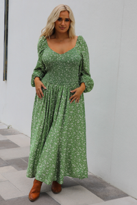 Woman model wearing a green maxi length dress that has a white floral print pattern, long sleeves, smocked bodice, and a flowy skirt. The model is holding the skirt up on one side. The photo is taken outdoors against a white wall. 