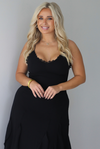 A woman wearing a black formal maxi dress with lace trim detailing at the bodice and ruffled details at the skirt, standing indoors against a white wall. The photo shows a closeup of the bodice of the dress, which has lace trim detailing at the neckline.