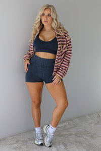 Woman wearing a pink, burgundy, lilac, and sand colored jacket with a charcoal colored two-piece set featuring a cropped sports bra and high waisted short biker shorts. The photo is taken indoors against a white wall with white marble flooring. 