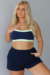 A blonde woman wearing a navy blue two-piece lounge set with light green trim, consisting of a sports bra and sweat shorts, standing indoors against a white wall.