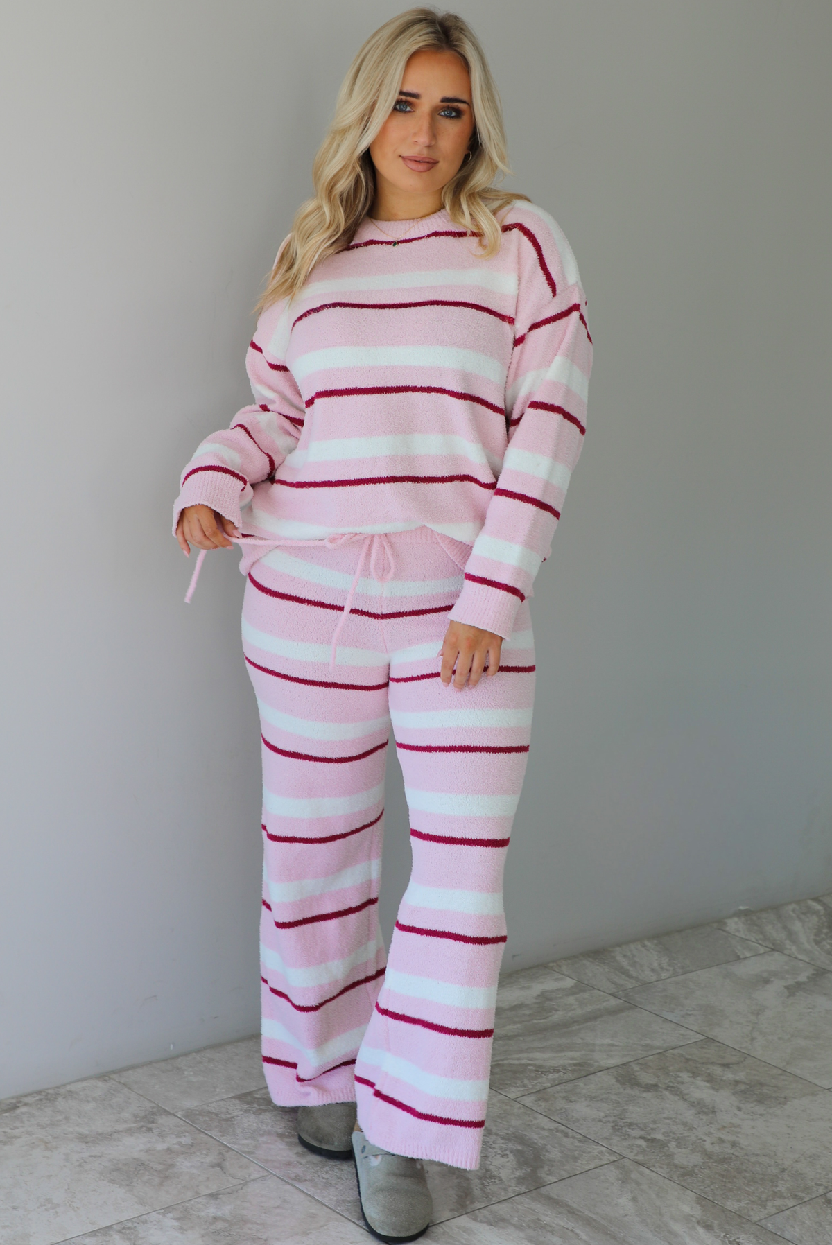 Woman wearing a pink set with a darker pink and white striped design. The set consists of an oversized sweater top and high waisted pants. The sweater of the set is tucked slightly in the front of the pants. The photo is taken indoors against a white wall with white marble flooring. 