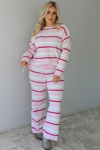 Woman wearing a pink set with a darker pink and white striped design. The set consists of an oversized sweater top and high waisted pants. The sweater of the set is tucked slightly in the front of the pants. The photo is taken indoors against a white wall with white marble flooring. 