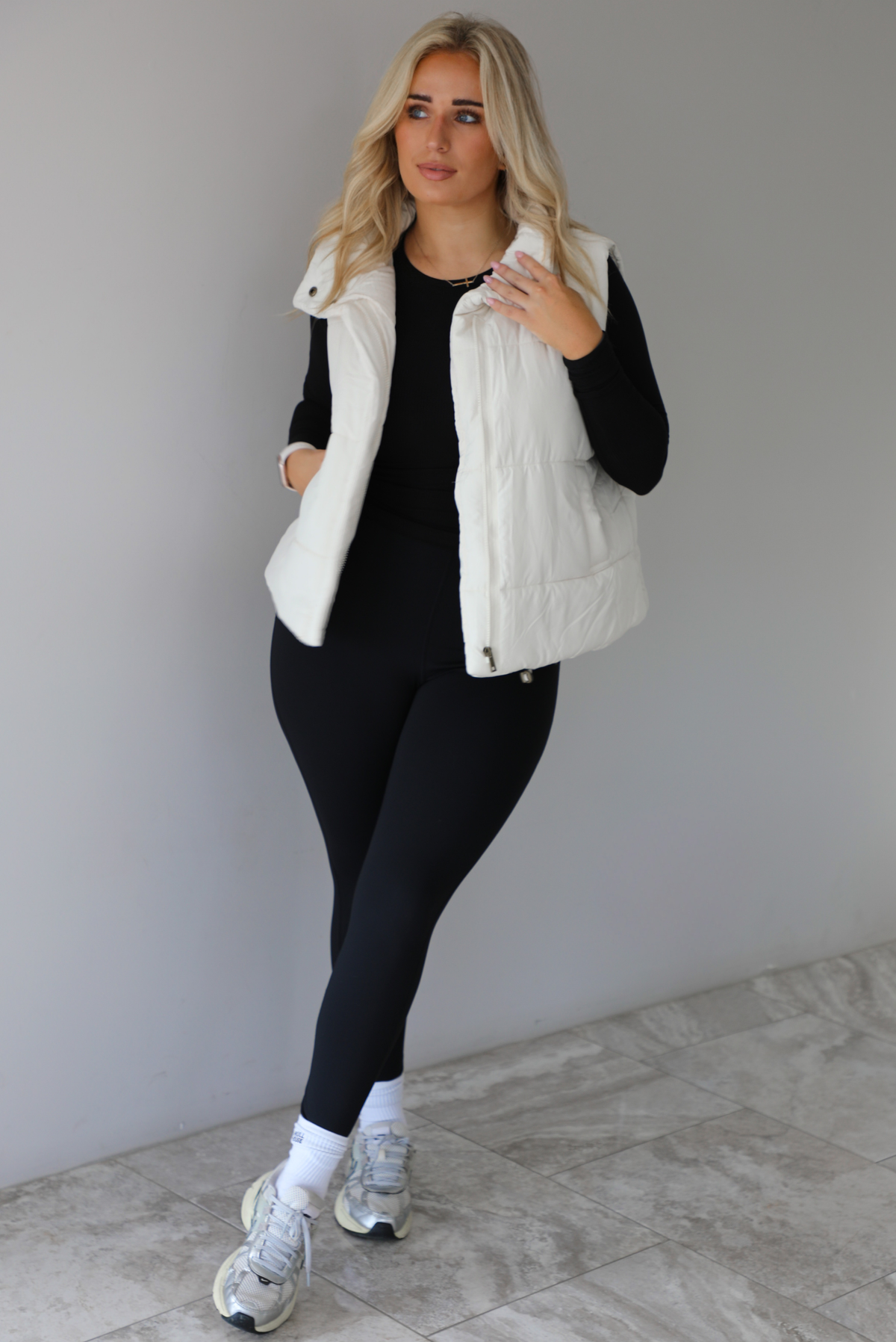 Woman wearing a light sand colored puffer vest with a fitted black long sleeved top and fitted leggings with high white ankle socks and silver sneakers. The photo is taken indoors against a white wall with white marble flooring. 